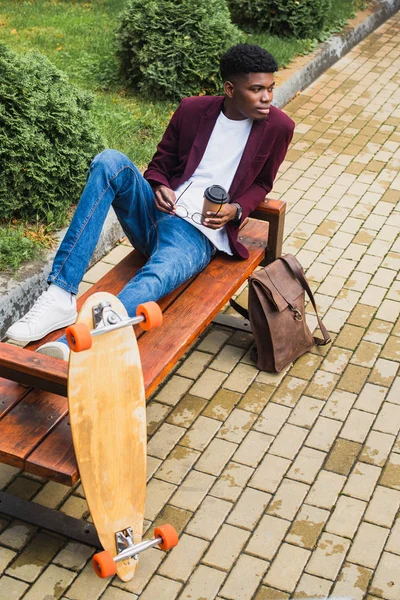 Joven con estilo con taza de papel de café y anteojos sentado en el banco - foto de stock