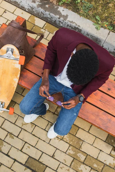 Вид сверху на молодого человека в очках и скейтборде, сидящего на скамейке — стоковое фото