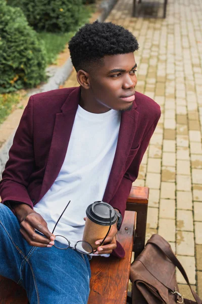 Красивый молодой человек с бумажной чашкой кофе и очками сидит на скамейке запасных — стоковое фото