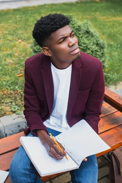 Jeune homme réfléchi assis sur le banc et écrit dans un carnet vierge — Photo de stock