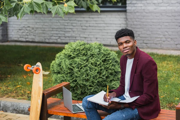 Jovem estudante afro-americano escrevendo em notebook na rua e olhando para a câmera — Fotografia de Stock