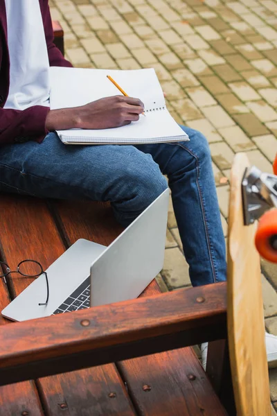 Colpo ritagliato di studente che scrive in notebook su panchina con computer portatile — Foto stock