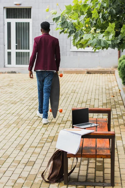 Visão traseira do homem andando com skate na rua com laptop e notebook no banco em primeiro plano — Fotografia de Stock