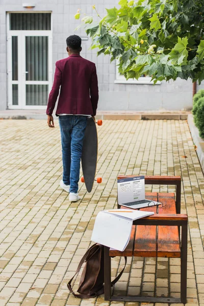 Задний вид фрилансера, идущего со скейтбордом на улице с ноутбуком с вэб-сайтом на экране, лежащим на переднем плане на скамейке — стоковое фото