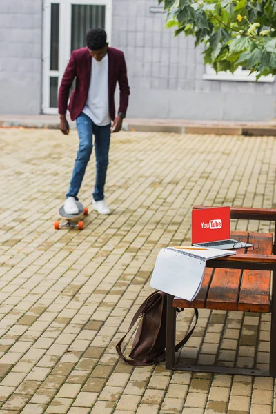 Young man riding skateboard on street with laptop with youtube logo on screen lying on bench on foreground — Stock Photo