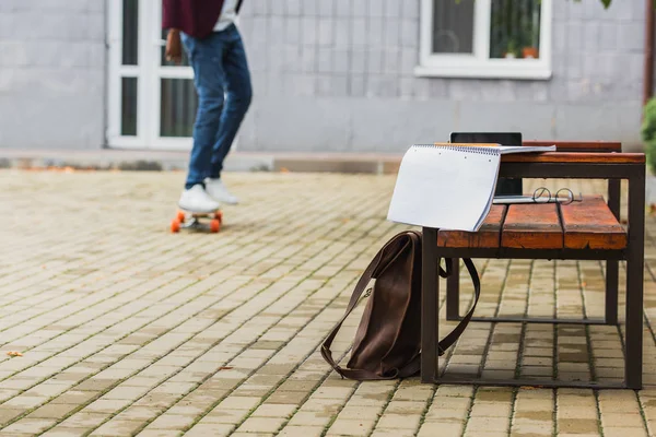 Cropped shot of student riding skateboard with backpack and notebook on bench on foreground — Stock Photo