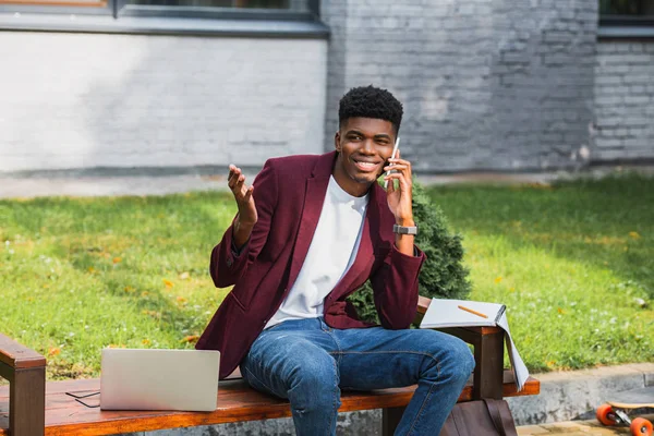 Smiling young freelancer talking by phone on bench with laptop — Stock Photo