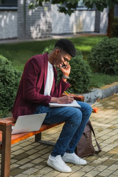 Souriant jeune pigiste parler par téléphone et écrire dans un cahier sur banc de rue — Photo de stock