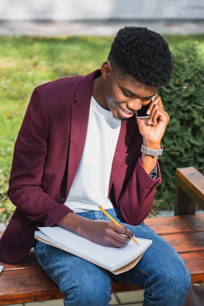 Jeune étudiant souriant parlant par téléphone et écrivant dans un cahier assis sur un banc — Photo de stock