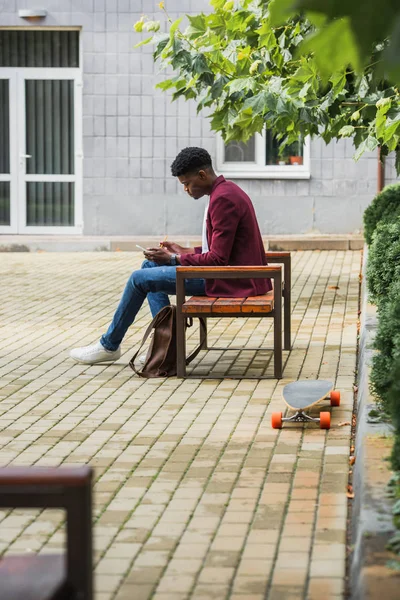 Стильний молодий студент, використовуючи смартфон, сидячи на лавці — стокове фото