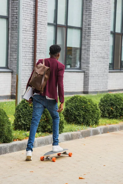 Visão traseira do estudante elegante com mochila e cadernos montando no skate na rua — Fotografia de Stock