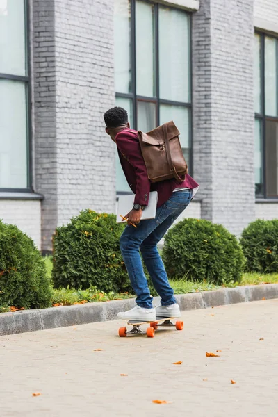 Rear view of young student with backpack and notebooks riding on skateboard on street — Stock Photo