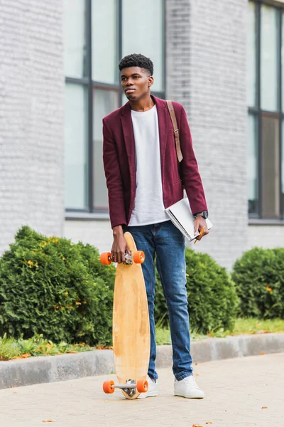 Jeune étudiant avec skateboard et cahiers debout dans la rue — Photo de stock