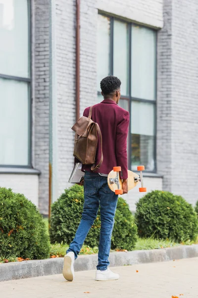 Vista posteriore del giovane studente con zaino e skateboard passeggiando per strada — Foto stock