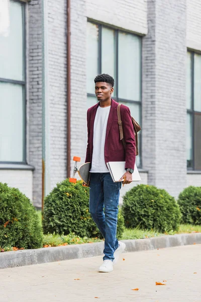 Joven estudiante guapo con mochila y monopatín caminando por la calle - foto de stock