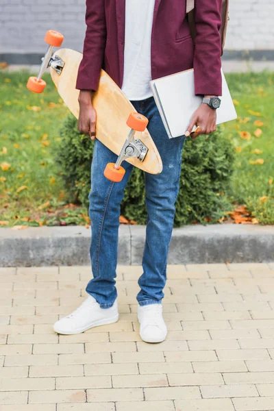 Cropped shot of student with notebooks and skateboard standing on street — Stock Photo