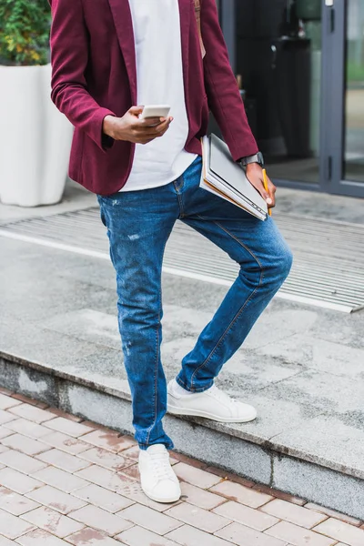 Cropped shot of stylish student with notebooks and smartphone standing on street — Stock Photo