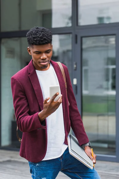 Handsome young student with notebooks and smartphone standing on street — Stock Photo