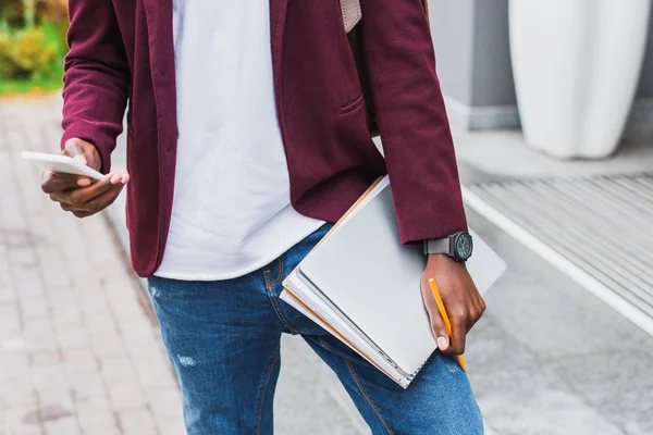 Cropped shot of student with notebooks and smartphone standing on street — Stock Photo