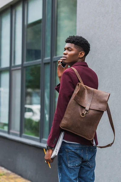 Handsome young student with leather backpack talking by phone on street — Stock Photo