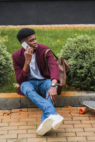 Handsome young man talking by phone while sitting on curb on street — Stock Photo