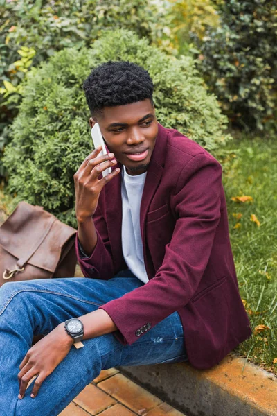 Smiling young man talking by phone while sitting on curb on street — Stock Photo