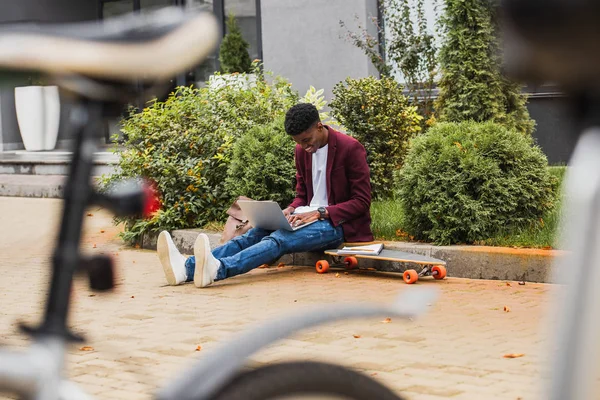 Young student using laptop and writing in notebook while sitting on curb on street — Stock Photo