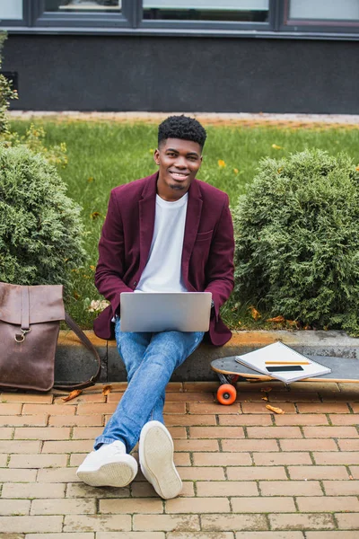 Щасливий молодий студент, використовуючи ноутбук, сидячи на бордюрі на вулиці і дивлячись на камеру — стокове фото
