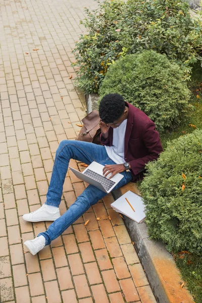 Jeune freelance travailler avec ordinateur portable et parler par téléphone tout en étant assis sur le trottoir sur la rue — Photo de stock