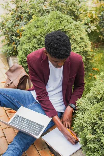 Vista de ángulo alto del joven freelancer utilizando el ordenador portátil y la escritura en el cuaderno mientras está sentado en la acera en la calle - foto de stock