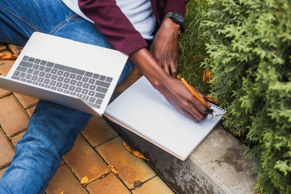 Cropped shot of freelancer using laptop and writing in notebook while sitting on curb on street — Stock Photo