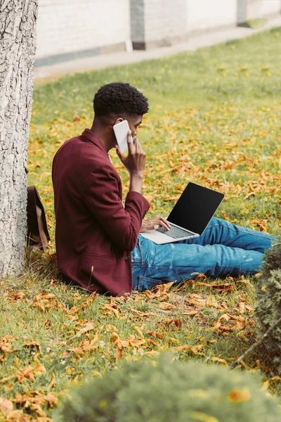 Афроамериканський фрілансер, що працює з ноутбуком і смартфоном на траві в парку — стокове фото
