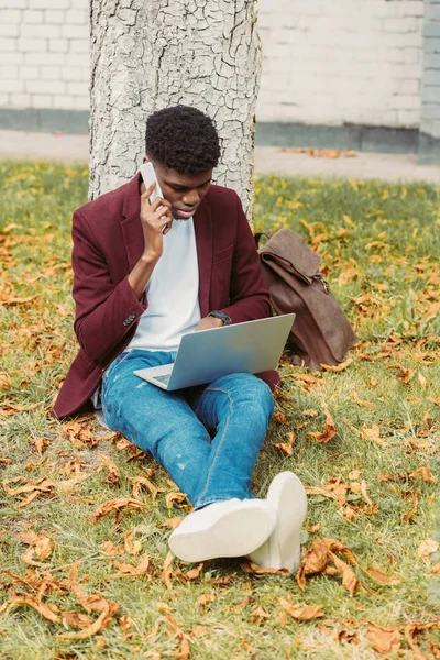 Guapo afroamericano freelancer trabajando con el ordenador portátil y teléfono inteligente en el parque - foto de stock
