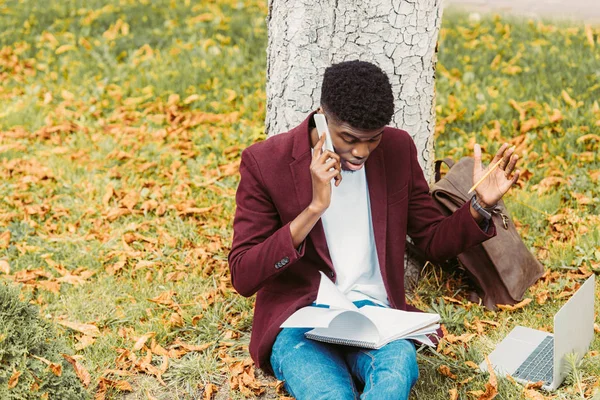 Confused african american freelancer working with laptop and talking on smartphone on grass in park — Stock Photo