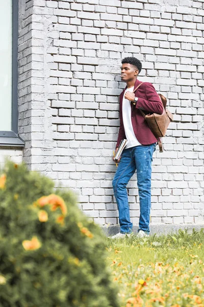 Étudiant afro-américain avec des documents et sac à dos en cuir debout près du mur de briques blanches — Photo de stock