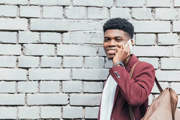 Homem americano africano alegre falando no smartphone perto da parede de tijolo branco — Fotografia de Stock