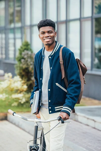 Beau Afro-Américain joyeux avec des documents et vélo dans la rue — Photo de stock