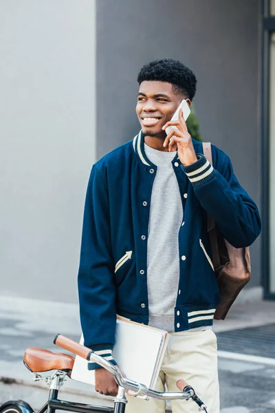 Bonito sorridente afro-americano homem com documentos falando no smartphone perto de bicicleta — Fotografia de Stock