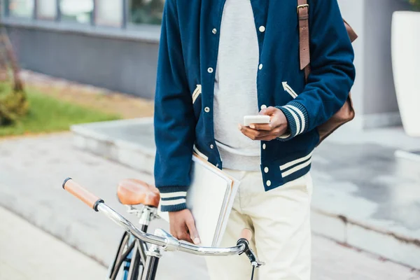 Vista cortada de teleworker americano africano elegante com documentos usando smartphone e de pé perto da bicicleta — Fotografia de Stock