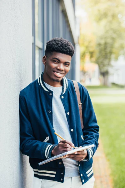 Alegre elegante africano americano estudiante escritura en libro de texto - foto de stock