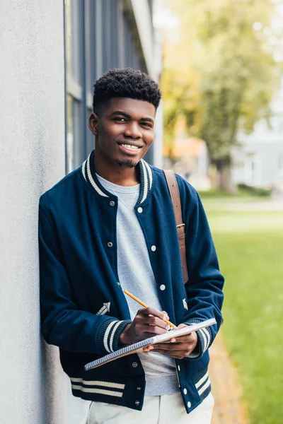 Sourire jeune étudiant afro-américain écrivant dans un manuel — Photo de stock