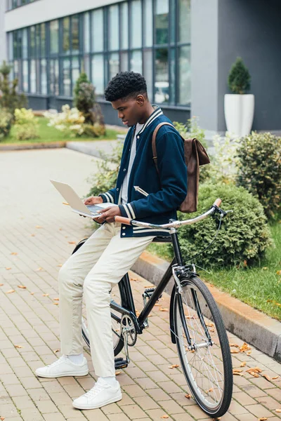 Hombre afroamericano con estilo haciendo trabajo remoto en el ordenador portátil mientras se apoya en la bicicleta en la ciudad - foto de stock