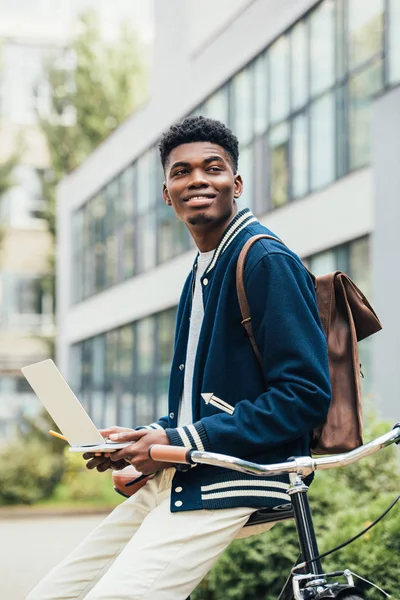 Alegre afroamericano teletrabajador utilizando portátil mientras se apoya en la bicicleta - foto de stock