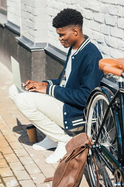 Elegante freelancer afroamericano que trabaja con el ordenador portátil cerca de la bicicleta con la mochila de cuero en la ciudad - foto de stock