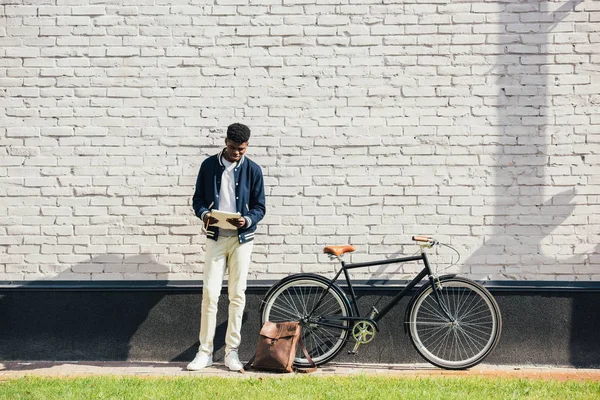 Freelancer afroamericano leyendo documentos y de pie cerca de la bicicleta en la pared de ladrillo blanco - foto de stock