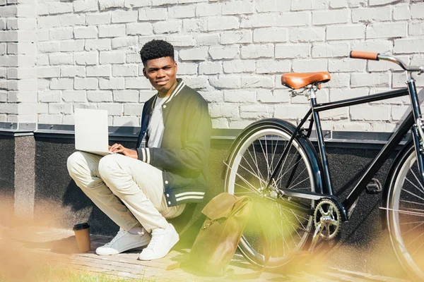 African american teleworker using laptop near bicycle at white brick wall — Stock Photo