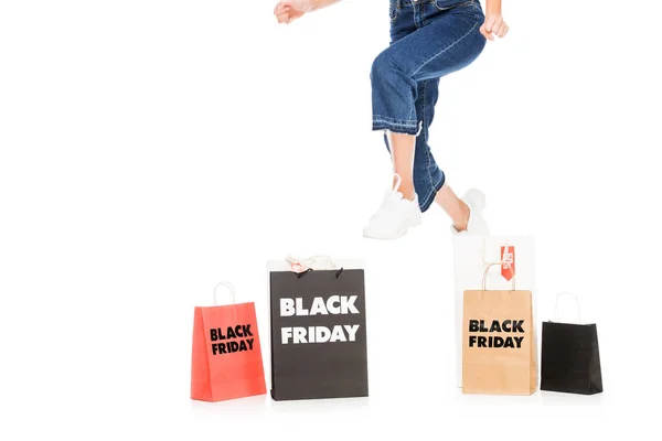 Vista recortada de niña saltando cerca de bolsas de compras con letreros de venta de viernes negro aislados en blanco - foto de stock