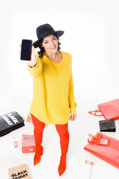 Приваблива модна дівчина, що показує смартфон з порожнім екраном біля сумки для покупок для чорної п'ятниці ізольовано на білому — стокове фото