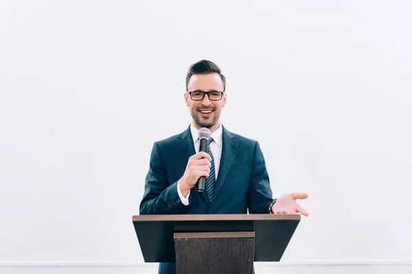 Smiling speaker gesturing and talking into microphone at podium tribune during seminar in conference hall — Stock Photo