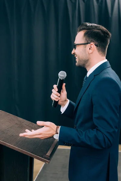 Vue latérale du haut-parleur gesticulant et parlant dans le microphone au podium tribune pendant le séminaire dans la salle de conférence — Photo de stock